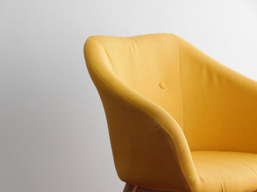 A person using a handheld vacuum cleaner to clean a decorative yellow and white patterned cushion placed on a dark fabric sofa in a residential living room, with a plain light-colored wall in the back