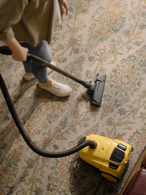 Close-up of a vacuum cleaner head positioned on a patterned, beige and pastel-colored carpet in a well-lit room. The vacuum attachment appears clean and functional, with visible control buttons. Part 