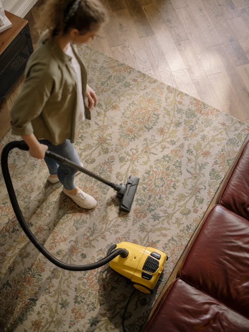A woman vacuuming a patterned, beige and pastel-colored rug in a living room with wooden floors and a brown leather sofa, demonstrating surface cleaning and maintenance for period flats. She is dresse
